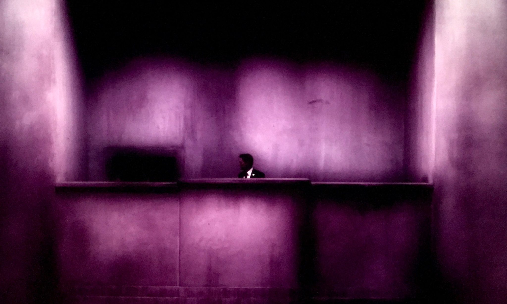 A lone figure in a dark suit behind a long counter, in a concrete hall washed in mauve light